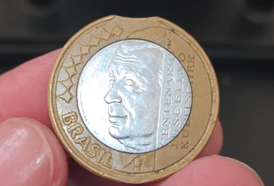 Moeda de 1 Real comemorativa de Juscelino Kubitschek (JK) sendo segurada, mostrando o raro erro de cunhagem "disco cortado" na parte superior, que a valoriza.