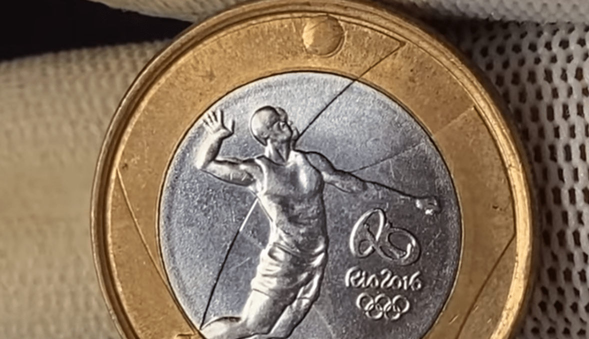 Close-up detalhado da moeda de 1 Real comemorativa do Vôlei (Rio 2016). A imagem foca no erro de duplicação, conhecido como 'cabeça dupla', que a torna uma moeda rara de alto valor.