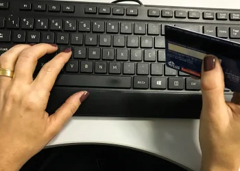 Mãos femininas digitando no teclado enquanto seguram um cartão de crédito azul para compras online.