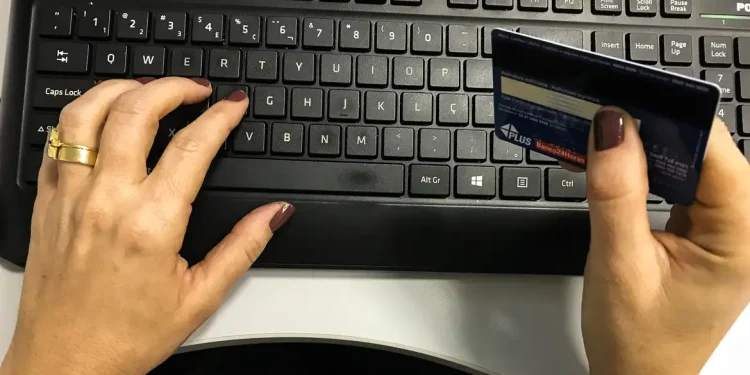 Mãos femininas digitando no teclado enquanto seguram um cartão de crédito azul para compras online.