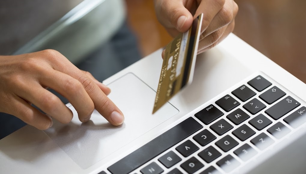 Mãos femininas digitando no teclado enquanto seguram um cartão de crédito azul para compras online.