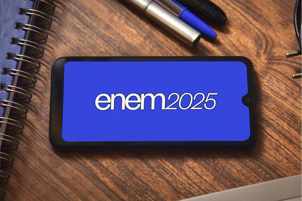 Smartphone sobre uma mesa de madeira exibindo a logo "ENEM 2025" em branco em uma tela azul. O celular simboliza a consulta digital do Cartão de Confirmação para verificar o local de prova.