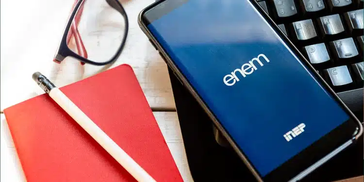 Imagem de tela de celular mostrando o logo do ENEM e Inep, sobre um teclado de notebook, junto a um lápis, caderno vermelho e óculos. Representa o momento em que o estudante confere seu local de prova e cartão de confirmação para o Enem 2025.