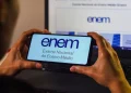 Duas mãos segurando um smartphone com o logo do ENEM (Exame Nacional do Ensino Médio) e a frase "Exame Nacional do Ensino Médio" na tela, enquanto um notebook ao fundo também exibe o logo do ENEM. A imagem ilustra o momento de solicitação de reaplicação do exame.