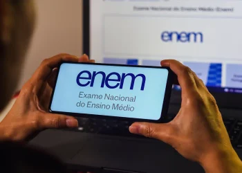 Duas mãos segurando um smartphone com o logo do ENEM (Exame Nacional do Ensino Médio) e a frase "Exame Nacional do Ensino Médio" na tela, enquanto um notebook ao fundo também exibe o logo do ENEM. A imagem ilustra o momento de solicitação de reaplicação do exame.
