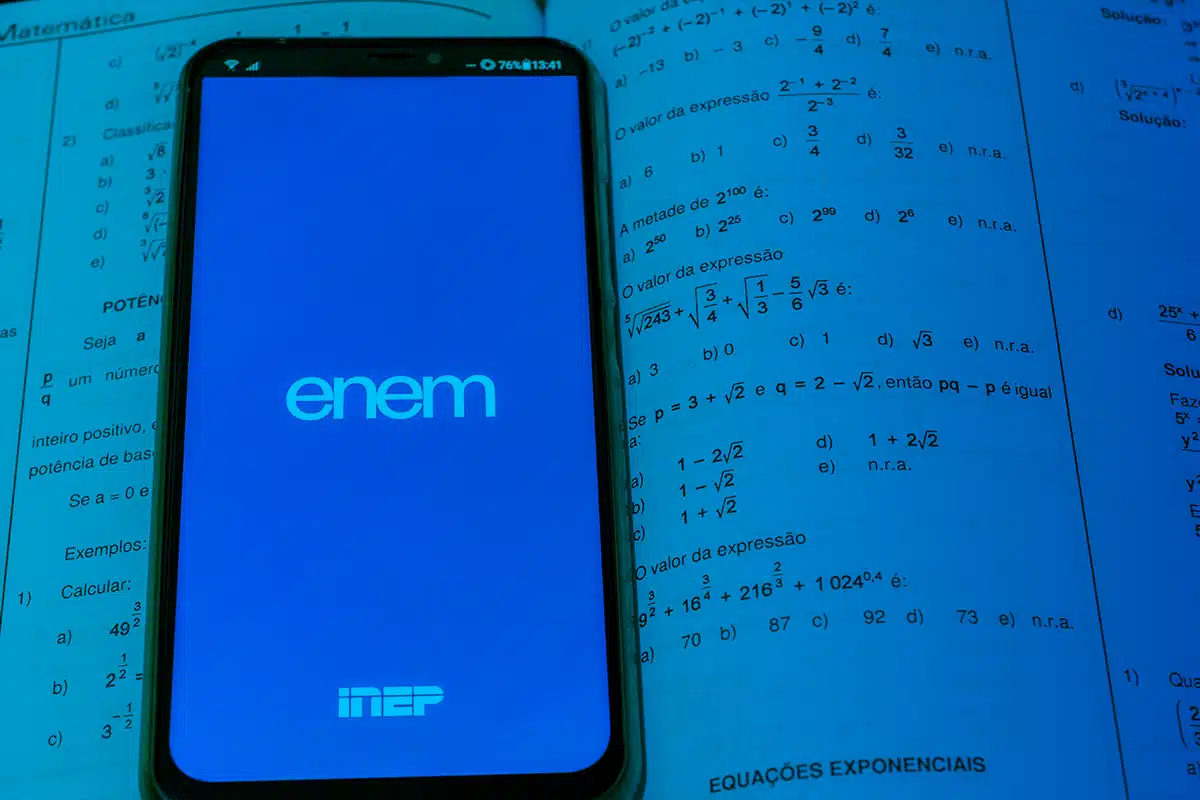 Celular mostrando tela do aplicativo ENEM aberto sobre apostila de matemática com exercícios e fórmulas.