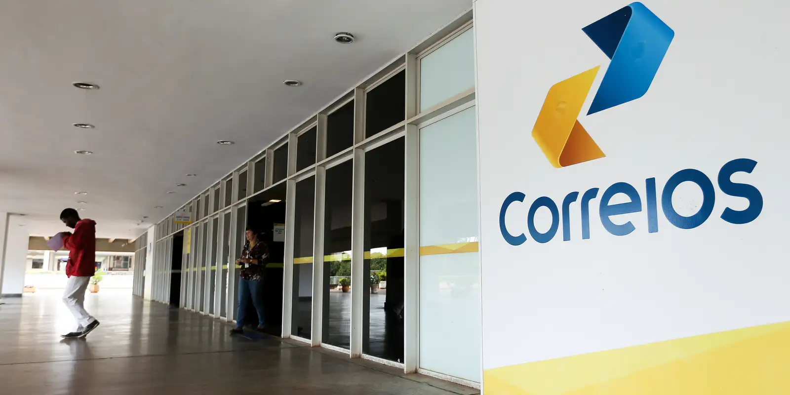 Fachada da agência dos Correios com logo colorido e pessoas dentro e fora do prédio