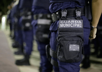 Uniforme da Guarda Municipal com bolsa preta e placa em destaque durante formação noturna