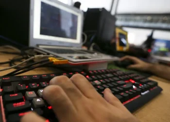 Mão digitando em teclado iluminado vermelho com laptop e equipamentos eletrônicos ao fundo