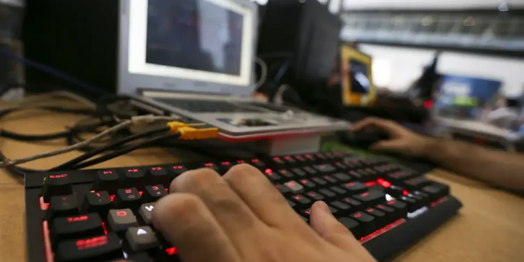 Mão digitando em teclado iluminado vermelho com laptop e equipamentos eletrônicos ao fundo