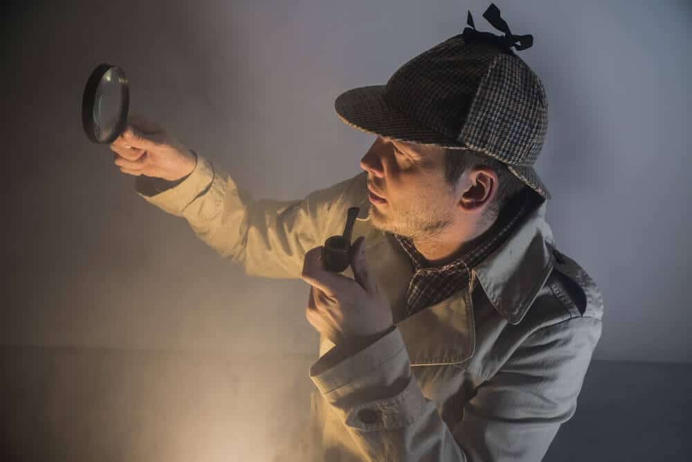 Detetive particular em ação, usando chapéu deerstalker e cachimbo, inspecionando uma pista com uma lupa em ambiente com névoa. Ilustra a profissão com salário médio de R$ 12.343.