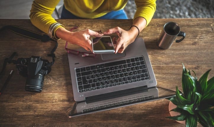 Visão aérea de uma mesa de madeira com uma pessoa trabalhando em um notebook. Estão presentes uma câmera profissional, um bloco de notas, um celular sendo segurado pelas mãos e uma caneca de metal.