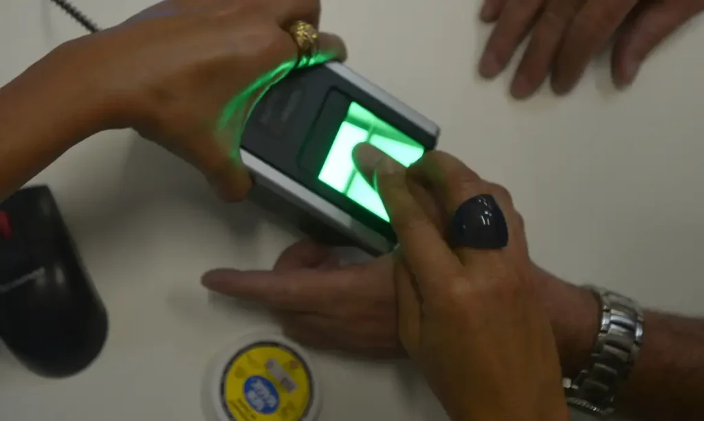Leitor de biometria verde capturando a impressão digital de uma pessoa com anéis, ilustrando a prova de vida ou cadastro biométrico para o INSS.