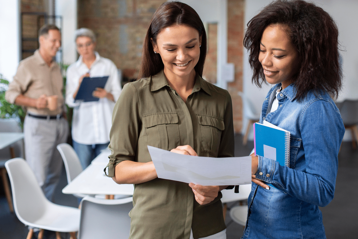 Duas mulheres sorrindo revisando documento em ambiente de trabalho com outras pessoas ao fundo.