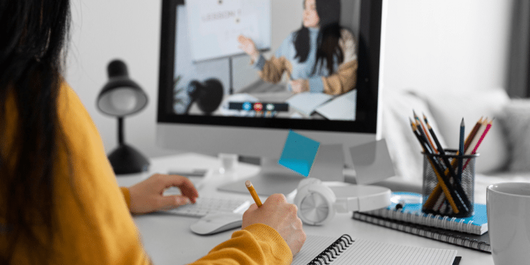 Pessoa realizando aula online no computador, anotando em caderno com lápis em mesa organizada