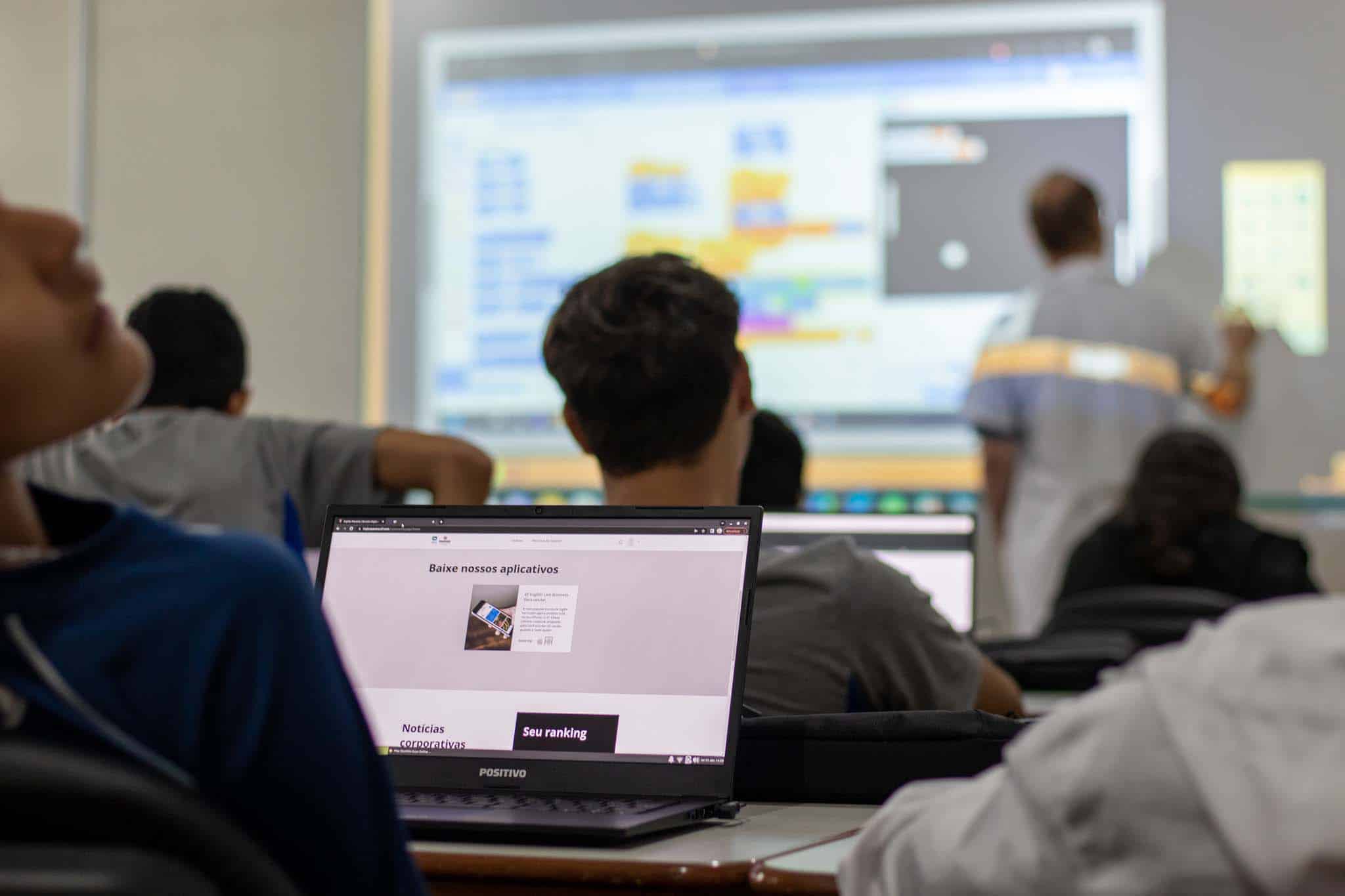 Alunos em sala de aula assistem professor que ensina programação com projetor enquanto usam notebooks