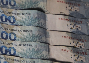 Pilha de cédulas de 100 reais emitidas pelo Banco Central do Brasil com design azul e detalhes visíveis.