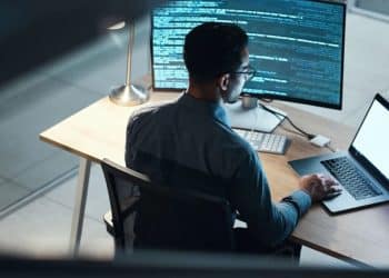 Programador focado trabalhando em código à noite com dois monitores, representando a busca por novas habilidades digitais e o avanço profissional em tecnologia.