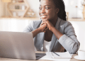 Mulher de negócios negra e sorridente, vestindo um blazer cinza claro, sentada em frente a um laptop prateado em um escritório doméstico bem iluminado. Ela apoia o queixo nas mãos, olhando para o lado, refletindo sobre estratégias de negócios e o futuro empreendedor.