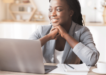 Mulher de negócios negra e sorridente, vestindo um blazer cinza claro, sentada em frente a um laptop prateado em um escritório doméstico bem iluminado. Ela apoia o queixo nas mãos, olhando para o lado, refletindo sobre estratégias de negócios e o futuro empreendedor.