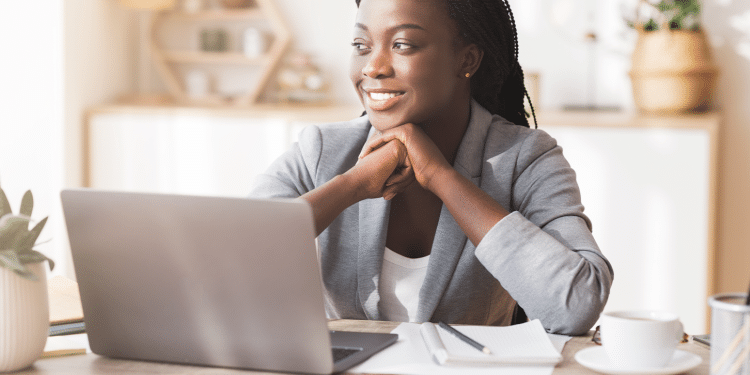 Mulher de negócios negra e sorridente, vestindo um blazer cinza claro, sentada em frente a um laptop prateado em um escritório doméstico bem iluminado. Ela apoia o queixo nas mãos, olhando para o lado, refletindo sobre estratégias de negócios e o futuro empreendedor.