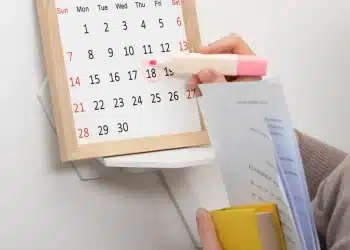 Mão marcando o dia 18 no calendário com marca-texto rosa segurando documentos e livro amarelo