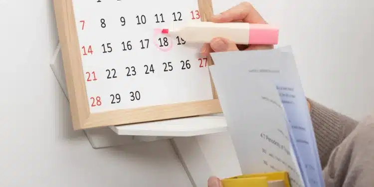 Mão marcando o dia 18 no calendário com marca-texto rosa segurando documentos e livro amarelo