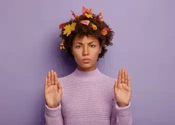 Pessoa com expressão séria fazendo gesto de pare, usando suéter lilás e adornos de folhas de outono no cabelo.