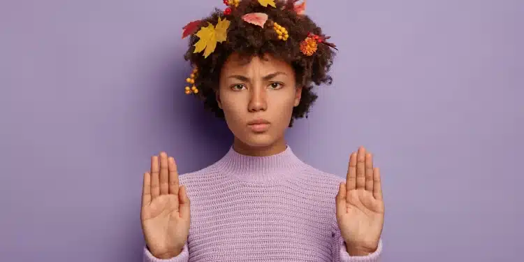 Pessoa com expressão séria fazendo gesto de pare, usando suéter lilás e adornos de folhas de outono no cabelo.