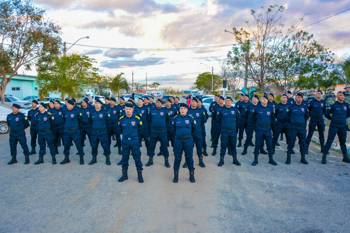Guardas Municipais uniformizados em formação, em fila, em área externa com carros estacionados ao fundo