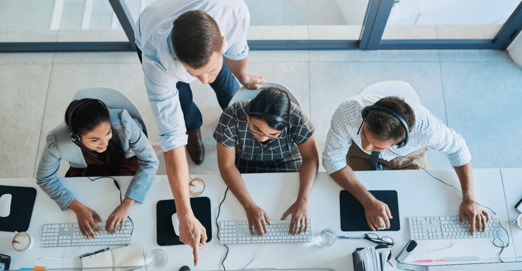 Vista superior de um gerente orientando uma equipe em call center ou escritório sobre o uso de habilidades digitais e tecnologia no serviço.