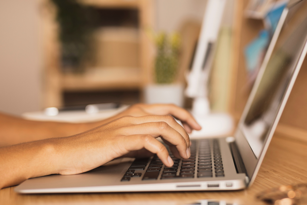 Mãos femininas digitando em teclado de notebook prateado sobre mesa de madeira clara em escritório.