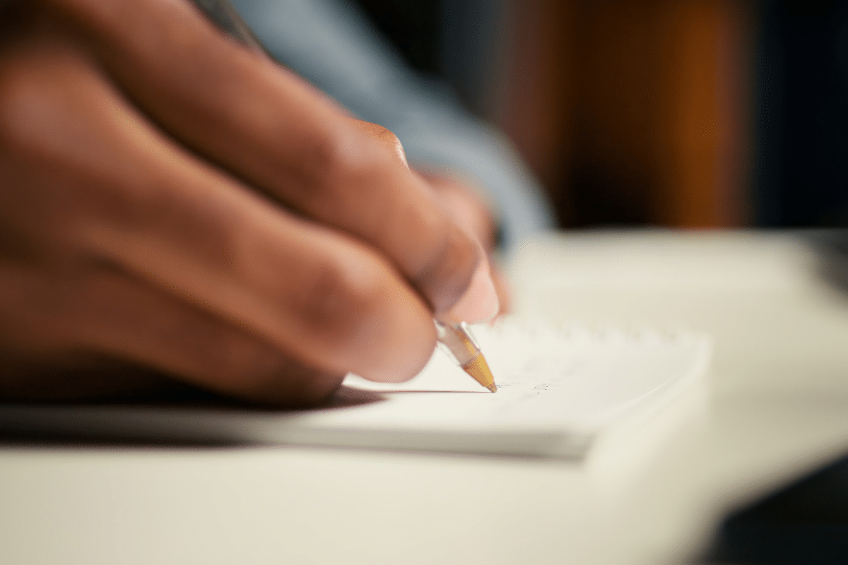 Pessoa segurando caneta e escrevendo em caderno branco sobre mesa clara