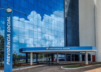 Fachada espelhada do prédio da Previdência Social com refletores azuis e céu com nuvens refletido.