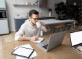Homem jovem e focado trabalhando remotamente em uma cozinha moderna, usando notebook e tablet, enquanto bebe café e anota em um caderno.