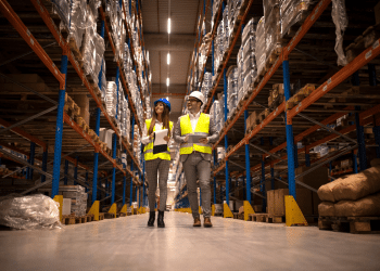 Gerentes de logística inspecionando corredor de armazenamento com mercadorias e equipamentos de segurança.