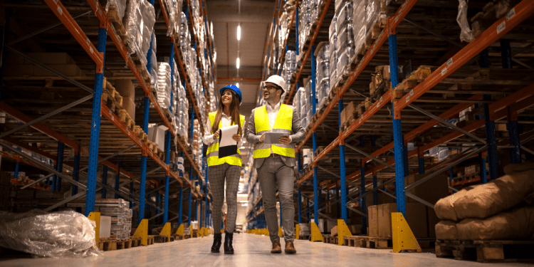 Gerentes de logística inspecionando corredor de armazenamento com mercadorias e equipamentos de segurança.