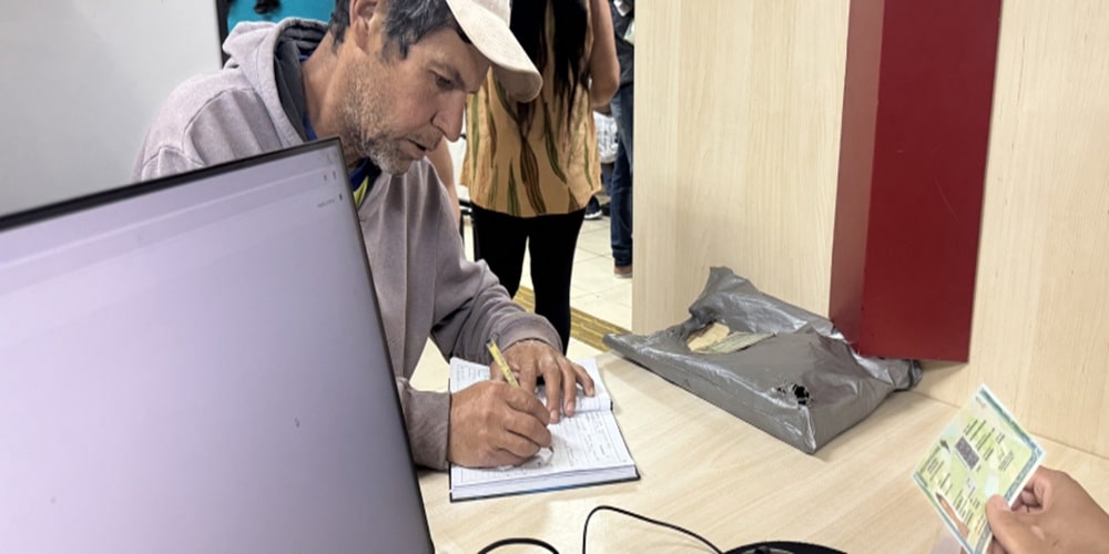 Homem preenchendo documento com caneta em balcão próximo a computador e documento em mãos.