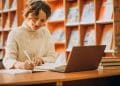 Estudante sorrindo e anotando em caderno enquanto usa notebook em mesa de biblioteca, com estantes de livros ao fundo.