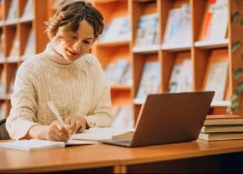 Estudante sorrindo e anotando em caderno enquanto usa notebook em mesa de biblioteca, com estantes de livros ao fundo.