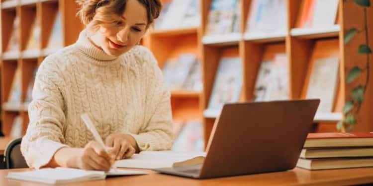 Estudante sorrindo e anotando em caderno enquanto usa notebook em mesa de biblioteca, com estantes de livros ao fundo.