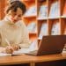 Estudante sorrindo e anotando em caderno enquanto usa notebook em mesa de biblioteca, com estantes de livros ao fundo.