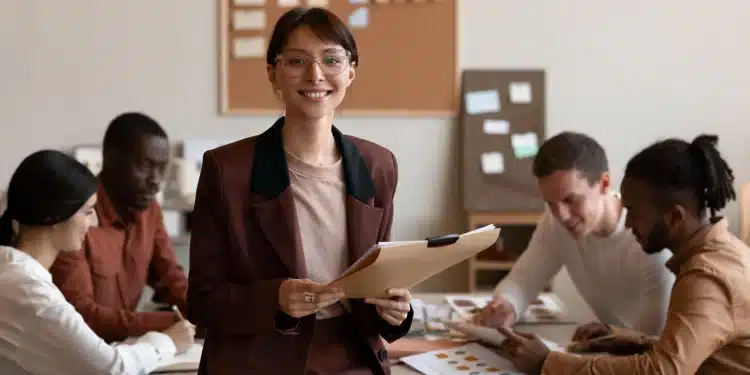 Mulher sorridente usando óculos e terno segurando pasta enquanto equipe diversa trabalha em mesa ao fundo
