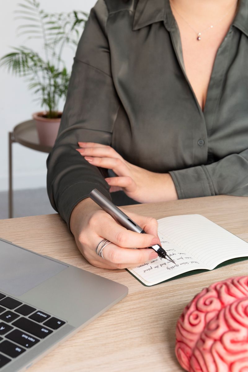 Profissional de saúde (psicólogo) fazendo anotações em um caderno, com um laptop aberto e um modelo de cérebro ao lado, simbolizando a sessão de terapia online e o estudo da mente.