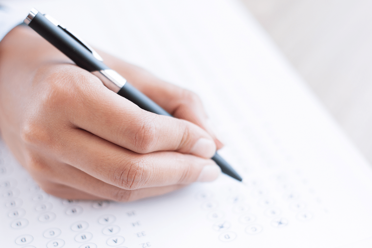 Mão segurando caneta preta preenchendo respostas em gabarito de múltipla escolha em papel branco