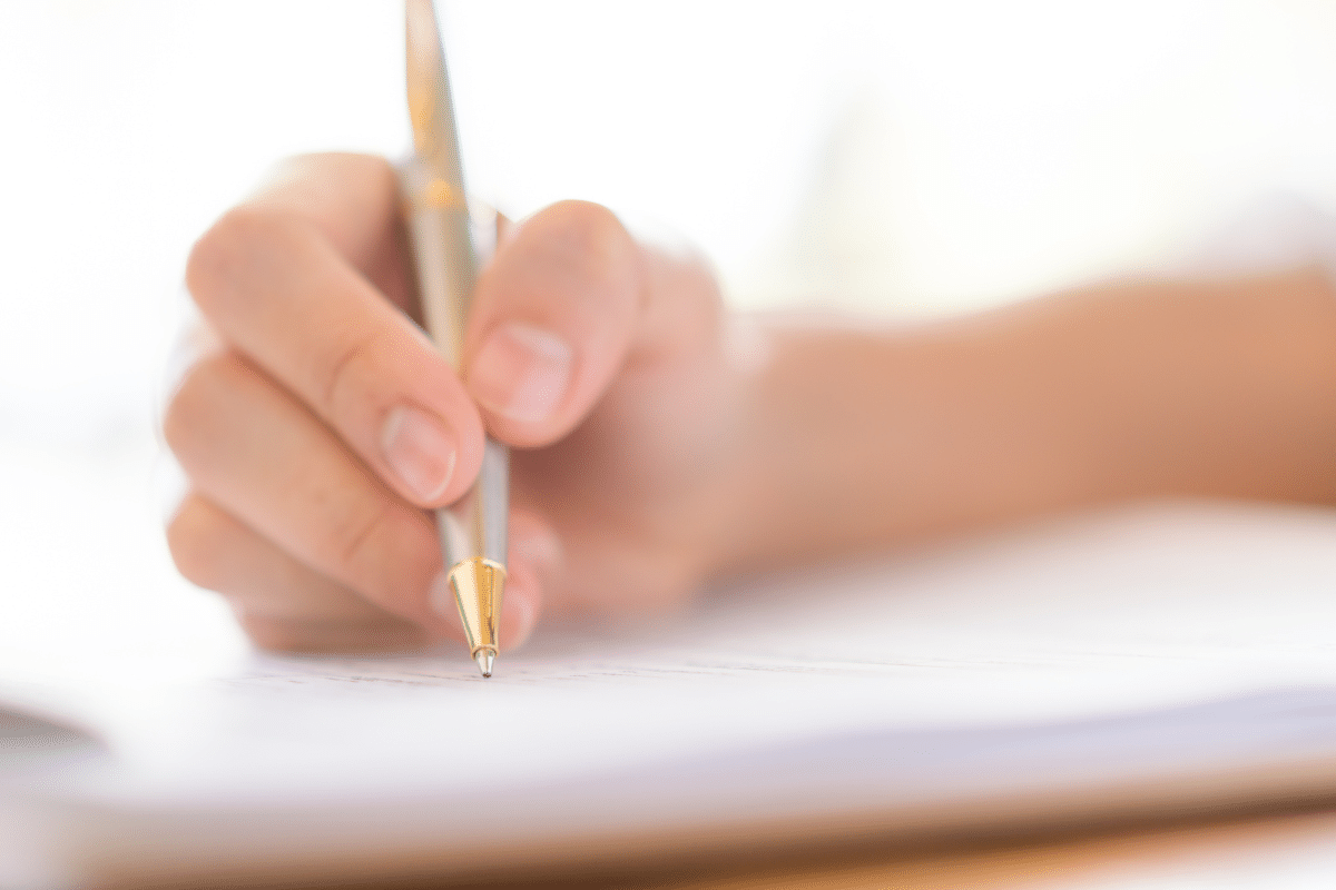 Mão segurando caneta metálica dourada escrevendo em papel branco desfocado ao fundo