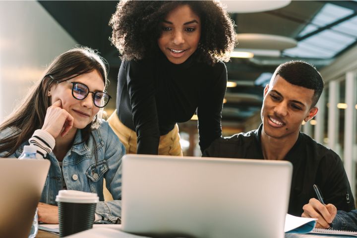 Três estudantes de diversas etnias colaboram olhando para um laptop em um ambiente moderno.