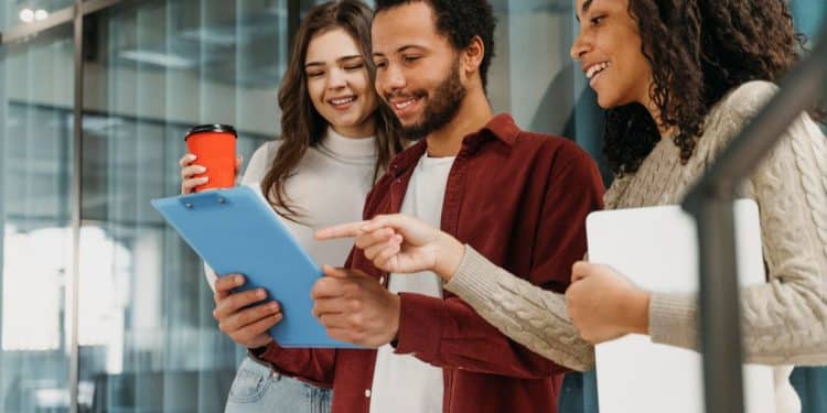 Três jovens profissionais em um escritório moderno discutindo um projeto em um clipboard. A imagem representa o planejamento de carreira e as profissões promissoras do futuro.