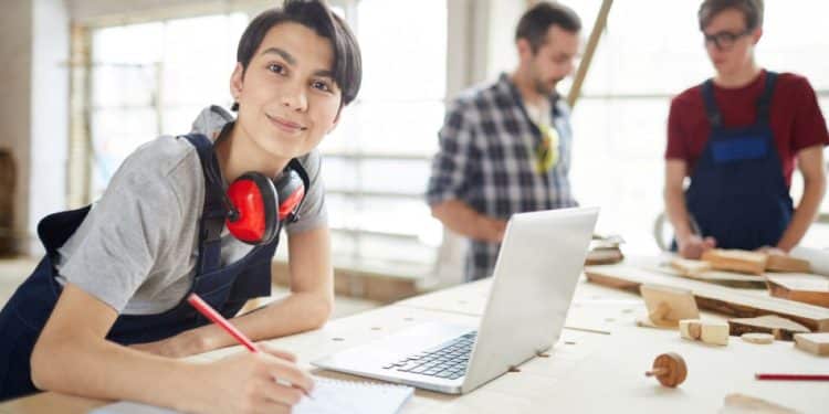 Jovem sorrindo anota em caderno, com fones de ouvido de proteção no pescoço. Pessoas desfocadas trabalham no fundo da oficina.