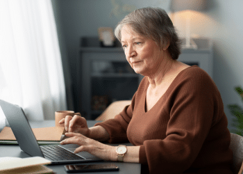 Mulher idosa com suéter marrom trabalhando e navegando em notebook próximo à janela iluminada